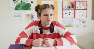 Sınıftaki genç kız sinirli görünüyor, kulaklık takıyor, masada oturuyor, okulda. Eğitim, teknoloji, gençlik, öğrenme, öğrenim, okul