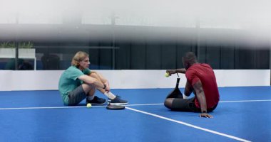 Diverse men sitting on padel indoor court, discussing game strategy with rackets in hand, copy space. teamwork, sports, competition, coaching, male, friendship