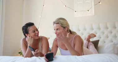 İki farklı bayan arkadaş yatakta dinleniyor, gülümsüyor ve evde akıllı telefon kullanıyor. Rahatlama, teknoloji, arkadaşlık, rahatlık, iletişim, yaşam tarzı