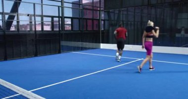 Genç yetişkinler mavi sahada Padel tenis oynuyorlar, rekabetin tadını çıkarıyorlar. Spor, raket, takım çalışması, rekabet, eğlence, atletizm