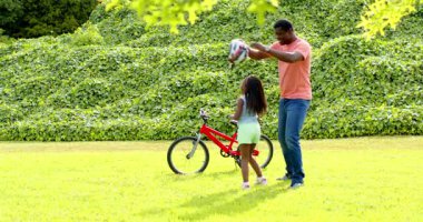 Afro-Amerikan, baba kızına güneşli parkta kırmızı bisiklete binmesinde yardım ediyor. Aile, ebeveynlik, açık hava, bağ, güvenlik, bisiklet