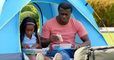 Afro-Amerikalı, baba-kız açık hava mavi kamp çadırında piknik yapıyorlar. Aile, bağ, doğa, rahatlama, yemek, eğlence.