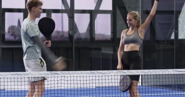 Genç adam ve kadın kapalı sahada Padel tenis oynuyorlar, tebessümlerle rekabetin tadını çıkarıyorlar. Spor, fitness, yarışma, raket, takım çalışması, egzersiz
