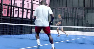 Padel tenisi oynayan iki farklı adam, kapalı sahada mavilikte yoğun bir şekilde odaklanıyor. Spor, atletizm, yarışma, takım çalışması, raket, oyun
