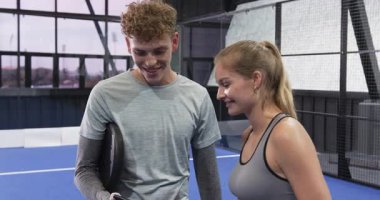 Genç adam ve kadın gülümsüyor, kapalı sahada Padel tenis maçından sonra akıllı telefonları kontrol ediyorlar. takım çalışması, teknoloji, iletişim, spor, ilişkiler, yaşam tarzı