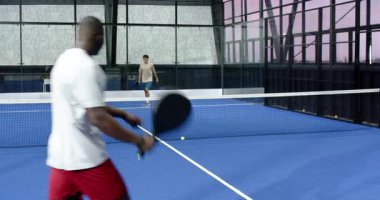 Kapalı sahada Padel tenis oynayan çeşitli adamlar, kararlılıkla maça odaklanıyorlar. Spor, raket, rekabet, takım çalışması, fiziksel aktivite, egzersiz