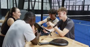 Farklı arkadaşlar kapalı sahada Padel tenis maçından sonra rahatlıyor, içki içiyor ve sohbet ediyorlar. Sosyalleşme, spor, eğlence, içecek, arkadaşlık, kutlama