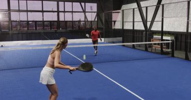Kapalı sahada Padel tenis oynayan iki farklı sporcu, yoğun ralliye odaklanıyor. Spor, rekabet, takım çalışması, çeşitli sporcular, aksiyon, raket