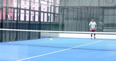Afrika kökenli Amerikalı bir adam kapalı sahada Padel tenisinde topu alıyor ve oynamaya hazırlanıyor. Spor, atletik, egzersiz, eğlence, rekabet, aktif yaşam tarzı