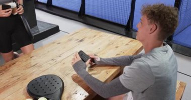 Ahşap masada oturan genç bir adam, elinde padel raketi ile, kapalı sahada maça hazırlanıyor. Spor, atlet, yarışma, ekipman, hazırlık, eğlence