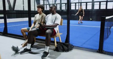 Çeşitli arkadaşlar yedek kulübesinde dinleniyor, Padel maçını izliyor, kapalı sahada boş vakitlerin tadını çıkarıyor. Rahatlama, dostluk, spor, seyircilik, dostluk, birliktelik.