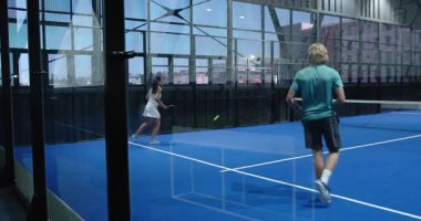 Kapalı alandaki mavi sahada Padel tenis oynayan çeşitli sporcular, takım çalışmasına odaklanıyorlar. Takım sporları, raket, işbirliği, fitness, rekabet, aktif