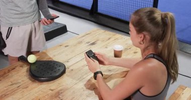 Bir kadın elinde kahveyle masada oturmuş, elinde padel raketi olan birinin içeri girişini izliyor. Tenis, rahatlama, rahatlama, yaşam tarzı, spor, dinlenme