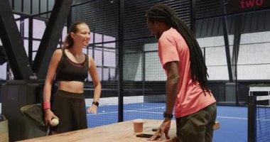 Padel tenis maçına hazırlanan çeşitli genç sporcular, kapalı sahada strateji tartışıyorlar. Takım çalışması, sportmenlik, rekabet, hazırlık, planlama