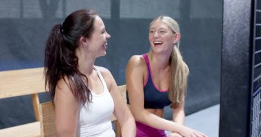 Spor kıyafetli kadınlar bankta oturur, kapalı sahada Padel tenis maçını dikkatle izlerler. İzleyiciler, aktif giyim, ilgi, beklenti, boş zaman, eğlence