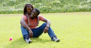 Afrika kökenli Amerikalı, baba ve kızı güneşli parkta çimlerin üzerinde birlikte gülüyorlar. Aile, bağ, mutluluk, açık hava, doğa, ebeveynlik