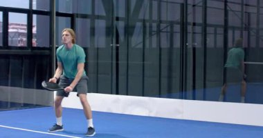 Kapalı alanda mavi kortta Padel tenis oynayan genç adam, odaklanmış ve harekete hazır. Spor, atlet, raket, rekabet, konsantrasyon
