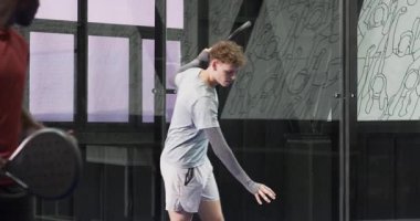 Padel raketi tutan genç bir adam, kapalı sahada güvenle gülümsüyor. Spor, atlet, raket, eğlence, egzersiz, fitness