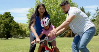 Ebeveynler kızlarına parkta bisiklet sürmeyi öğretiyor, gülümsüyor ve dışarıda eğleniyorlar. Aile, bağ, ebeveynlik, bisiklet, doğa, destek