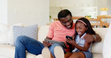 Afro-Amerikan, baba-kız tablet ve akıllı telefon kullanarak kanepede birlikte gülüyorlar. Aile, bağ, teknoloji, eğlence, oturma odası, ebeveynlik.