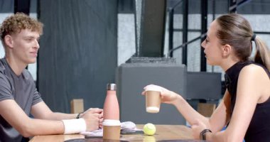 Young man smiling while sitting with friend, enjoying break after padel tennis, on indoor court. Friendship, leisure, sports, relaxation, laughter, recreation