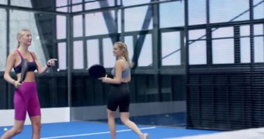 Kadınlar kapalı alanda mavi kortta Padel tenis oynuyorlar, oyun stratejilerine odaklanıyorlar. Spor, takım çalışması, sporcular, raket, rekabet, fitness