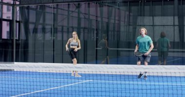 Genç yetişkinler kapalı alanda mavi sahada, topa odaklanarak Padel tenis oynuyorlar. Spor, rekabet, takım çalışması, raket, enerji, eğlence