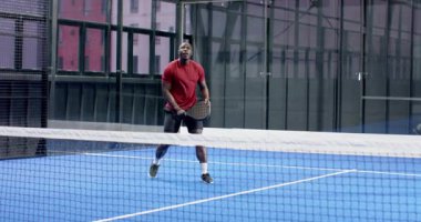 African American man playing padel tennis, hitting ball with focus and energy, at indoor court. sport, athlete, racquet, energetic, competition, active