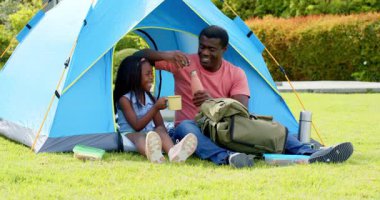 Afro-Amerikalı, baba-kız çadırın dışında piknik yapıyor, atıştırmalıkları ve kahkahaları paylaşıyorlar. Aile, bağ, açık hava, kamp, boş zaman, birliktelik.