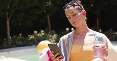Havuzda dinlenen, akıllı telefondan ve güneşli bir günde ferahlatıcı içkiden hoşlanan bir kadın. Boş zaman, tatil, teknoloji, yaz, güneş banyosu, havuz başı
