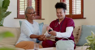 Önlüklü bir bakıcı evde yaşlı bir kadını rahatlatıyor, sıcak sohbetler ediyor. Bakıcı, şefkat, destek, hemşire, sağlık, bakım