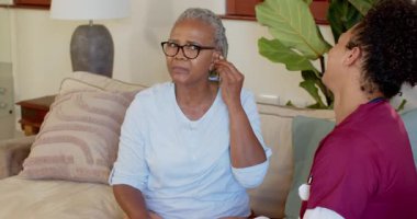 Kıdemli kadın işitme cihazını düzeltirken, önlüklü bakıcı evde yardım öneriyor. Yaşlılar, sağlık hizmetleri, tıbbi cihaz, destek, işitsel bilimler