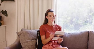 Kanepede dinlenen, kahvesini yudumlayan ve pencere kenarından kitap okuyan bir kadın. Rahatlama, rahatlama, yaşam tarzı, ev, konfor, hafta sonu