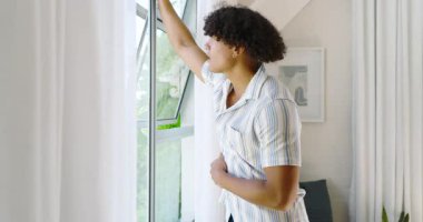 Çizgili tişörtlü, gülümseyen genç adam açık pencerenin yanında, evde temiz havanın tadını çıkarıyor. Mutluluk, rahatlama, özgürlük, günlük, yaşam tarzı, pozitif