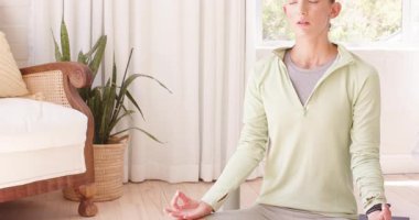 Kadın evde meditasyon yapıyor, pencerenin yanında gözleri kapalı, huzur içinde oturuyor. Meditasyon, sükunet, farkındalık, rahatlama, dinginlik, sağIık