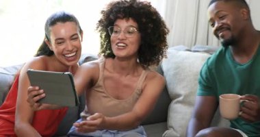 Çeşitli arkadaşlar kanepede birlikte gülüyor, evde tablet eğlencesinin tadını çıkarıyorlar. Teknoloji, arkadaşlık, rahatlama, gevşeme, bağlanma, alet