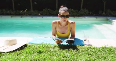 Havuz başında dinlenen, güneş gözlüklü bir kadın yanında canlandırıcı içecek olan kitap okuyor, fotokopi çekiyor. Tatil, tatil, rahatlama, havuz kenarı, yaz tatili, yaşam tarzı.
