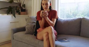 Kanepede dinlenen, elinde kahve fincanı tutan, evinde huzurlu bir anın tadını çıkaran bir kadın. Rahatlık, rahatlık, rahatlık, huzur, yaşam tarzı, sıcaklık.