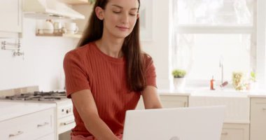 Mutfakta dizüstü bilgisayar kullanan gülümseyen kadın, sabah rutininin keyfini çıkarıyor. Teknoloji, yaşam tarzı, kahve, çalışma, çoklu görev, ev ofisi