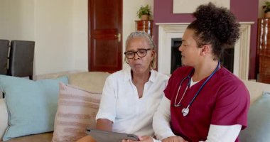 Tabletli yaşlı kadın oturma odasında hemşireyle konuşuyor, evde bakımı tartışıyor. Bakım, sağlık, iletişim, yaşlılık, hemşirelik, destek