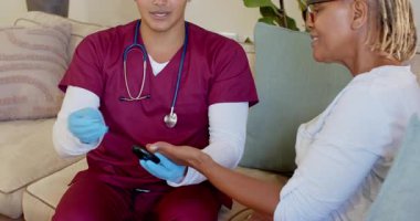 Önlüklü bir bakıcı, evde yaşlı bir kadına akıllı telefon gösteriyor, içten gülümsüyor. Bakım, sağlık, ev bakımı, arkadaşlık, yardım, hemşirelik