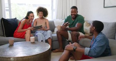 Çeşitli arkadaşlar kanepede gülüyor, sohbet ediyor, kahve ve tabletin tadını çıkarıyorlar. Arkadaşlık, eğlence, teknoloji, sohbet, bağ, rahatlama.