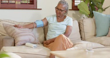 Evde kan basıncını ölçen yaşlı bir kadın, rahat bir koltukta oturuyor. Kıdemli, sağlık, sağlık, izleme, yaşam tarzı, rutin