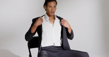 Asyalı erkek model çerçeveye giriyor, stüdyoda sandalyede oturuyor ceketi poz vermek için ayarlıyor. Profesyonel, minimalist, moda, iş, zarif, portre, stüdyo