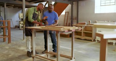African American mentor pointing guiding apprentice measuring wood with tape marking cuts at shop. Craftsmanship, woodworking, training, industrial, collaborative, rustic, skill