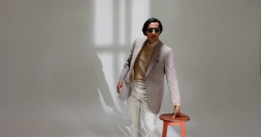 Asyalı erkek model pencere ışığında stüdyoya giriyor, ceketi ayarlıyor ve çeviriyor, turuncu taburede oturuyor. Moda, erkek, stüdyo, tarz, zarafet, modern, kendinden emin.