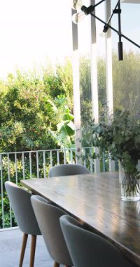 Dikey video: Büyük pencereli ahşap yemek masasının üzerinde cam vazoda şık bir çiçek aranjmanı. Dekor, iç mekan, tasarım, çiçek, süs, güneş ışığı