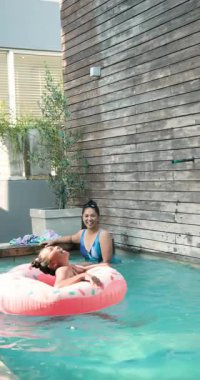 Dikey video: Anne ve kızı pembe çörek arabasıyla havuz başında eğleniyor, birlikte gülümsüyorlar. Aile, bağ, rahatlama, yaz, havuz başı, su.