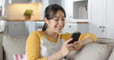 Telefon bildirimi alınıyor, Asyalı kadın gri kanepeye yaslanıyor, gülümsüyor ve cevap veriyor. Rahat, rahat, modern, yaşam tarzı, rahatlama, iletişim, ev