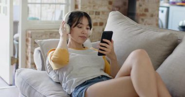 Müzik başlatmak için akıllı telefon ekranını dinlemek, Asyalı bir kadının kulaklık kaldırıp çatı katında yastığa uzanması. Rahatlama, rahatlama, modern yaşam tarzı, konfor, dinginlik, teknoloji